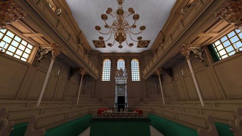 Digital image. A wood-panelled, hall-like room has three rows of seating along the three walls we see, and there is a balcony above. A brass chandelier with two levels of curving arms hangs from the ceiling. A chair at the far end is surrounded by elaborate moulding and ornament.