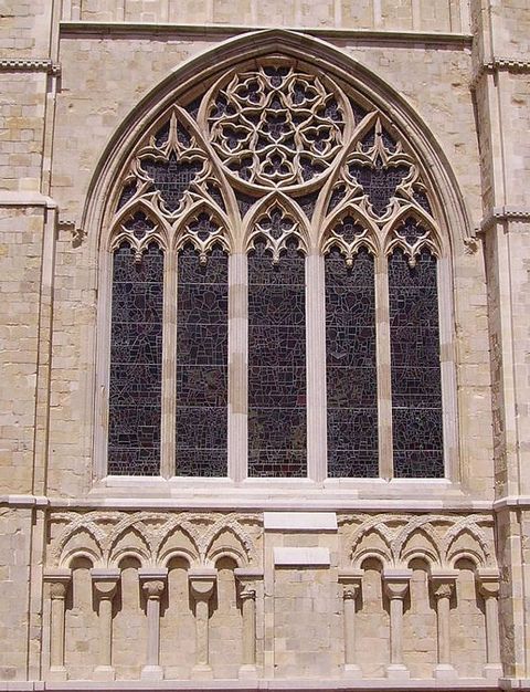 Architecture. From outside, we look up at a pointed arched stained glass window. The window is divided into five columns with a rose window at the top centre. Tracery gives the window a lacy look.