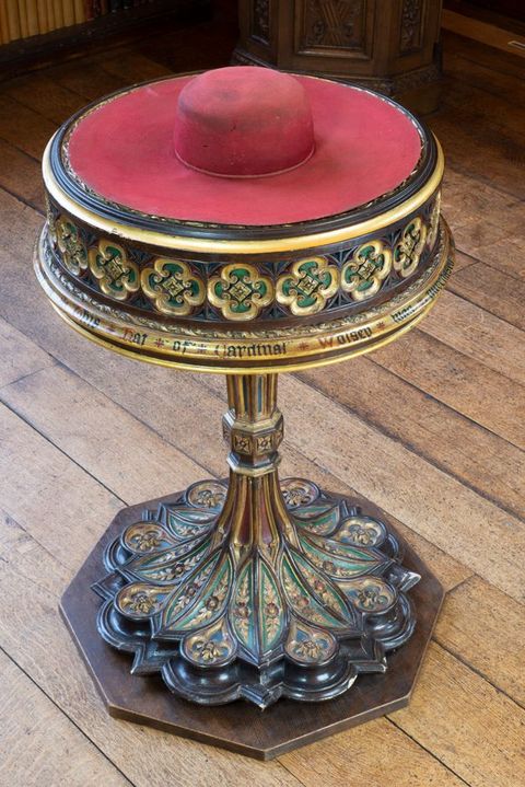 A red, wide-brimmed chat with a tall, rounded crown sits on an elaborately carved and painted plinth resembling a tall cake stand. Text around the gilded moulding at the bottom of the segment holding the hat reads, “Hat of Cardinal Wolsey”.