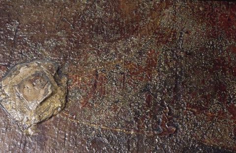 Painting. Detail view of a diamond-shaped object in low relief is gilded, as are thin lines emanating from it. The rest of the paint appears textured with burgundy red and grey.